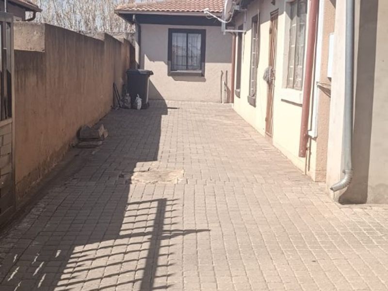 A paved side walkway along the exterior of the house, featuring boundary walls, drainage downpipes, and security bars on the windows.