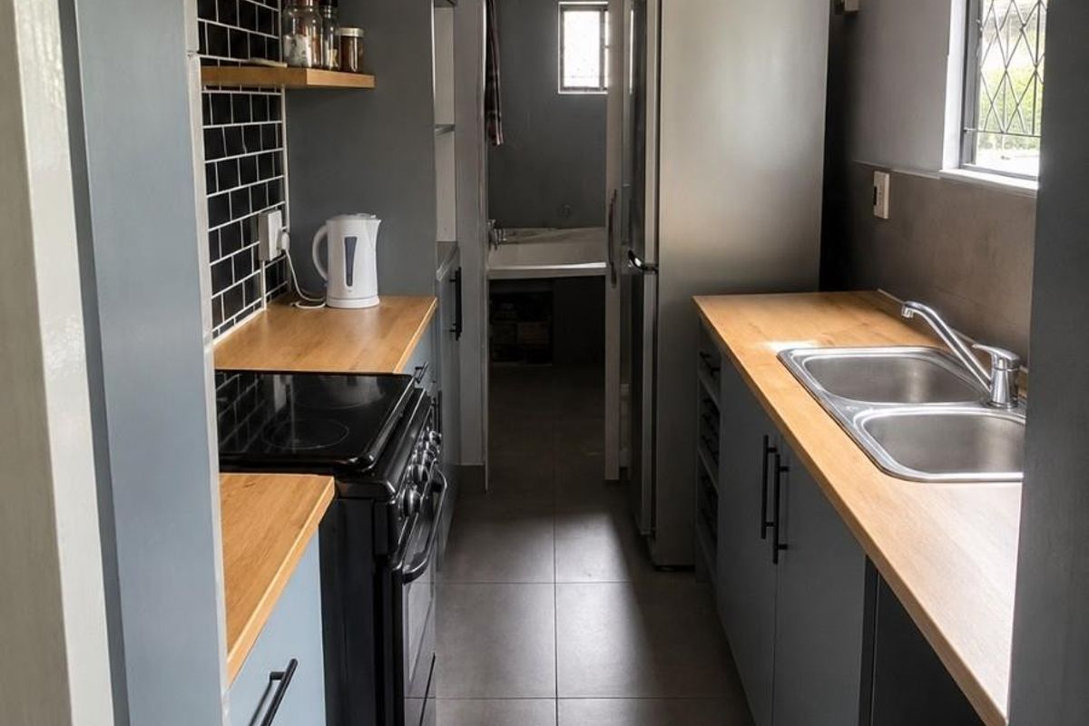 Modern galley-style kitchen featuring grey cabinetry, wooden countertops, black subway tile backsplash, and floating shelves, leading through to a secondary utility area.