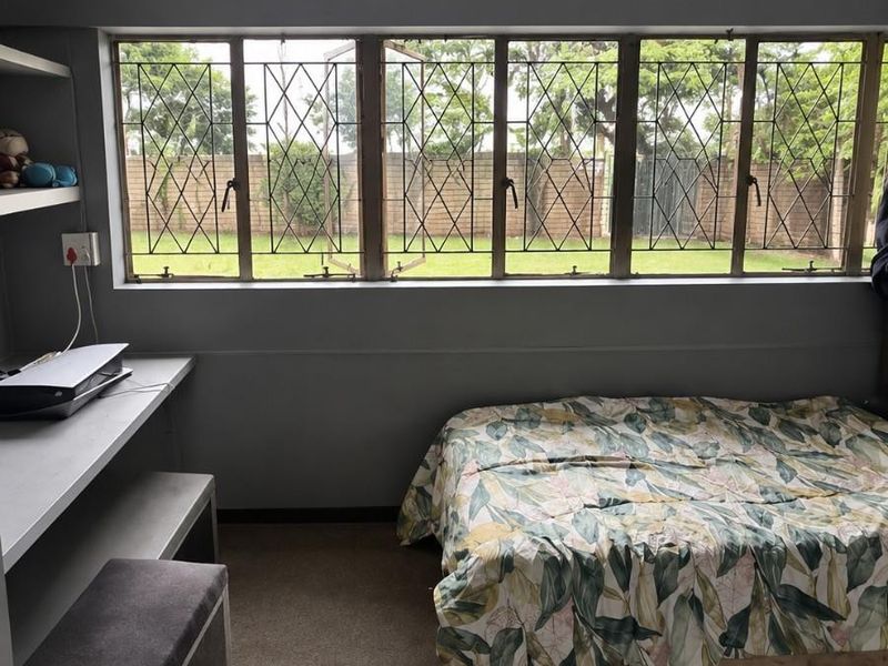 A bedroom featuring built-in desk and shelving, carpeted flooring, and large windows with security bars overlooking the rear garden.