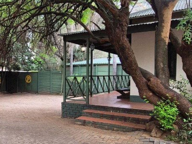 A charming front exterior view featuring a covered veranda with brick steps, timber railings, and a paved driveway shaded by a large mature tree.