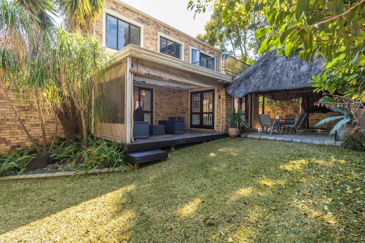 Rear view of the property featuring a brick facade, a covered timber deck patio, a traditional thatched lapa, and a landscaped garden.