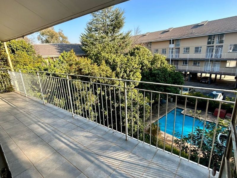 A spacious covered balcony featuring tiled flooring and a metal balustrade, offering a pleasant view over the complex garden and swimming pool area.