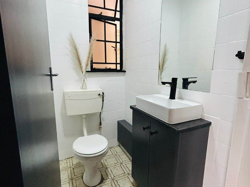 A compact bathroom featuring a modern vanity with a vessel sink, black fixtures, white wall tiling, and a window with a black frame.