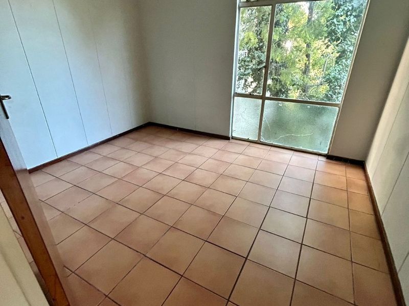 An empty bedroom featuring tiled flooring and a large window with a cracked pane, offering natural light and views of surrounding greenery.
