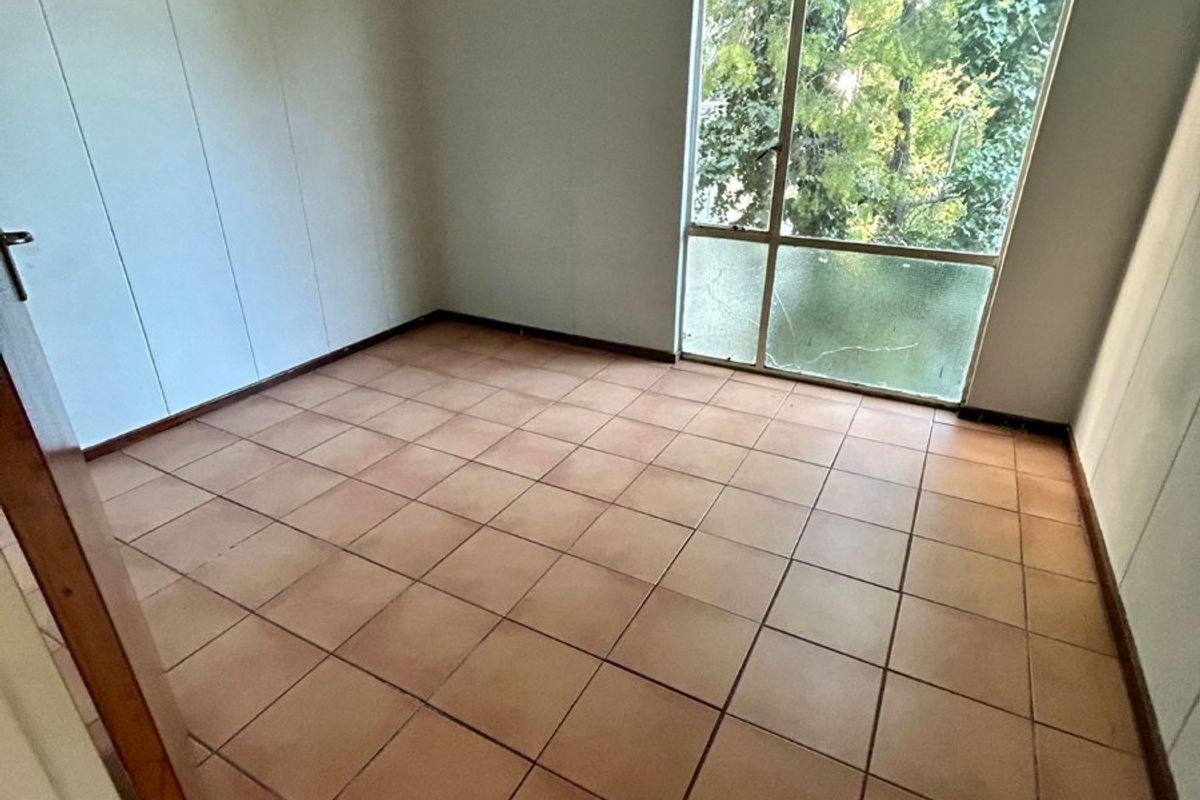 An empty bedroom featuring tiled flooring and a large window with a cracked pane, offering natural light and views of surrounding greenery.