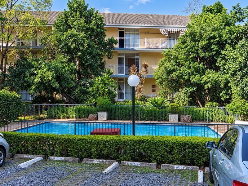 Exterior view of an apartment complex featuring a swimming pool, secure metal fencing, and private balconies overlooking the landscaped grounds.