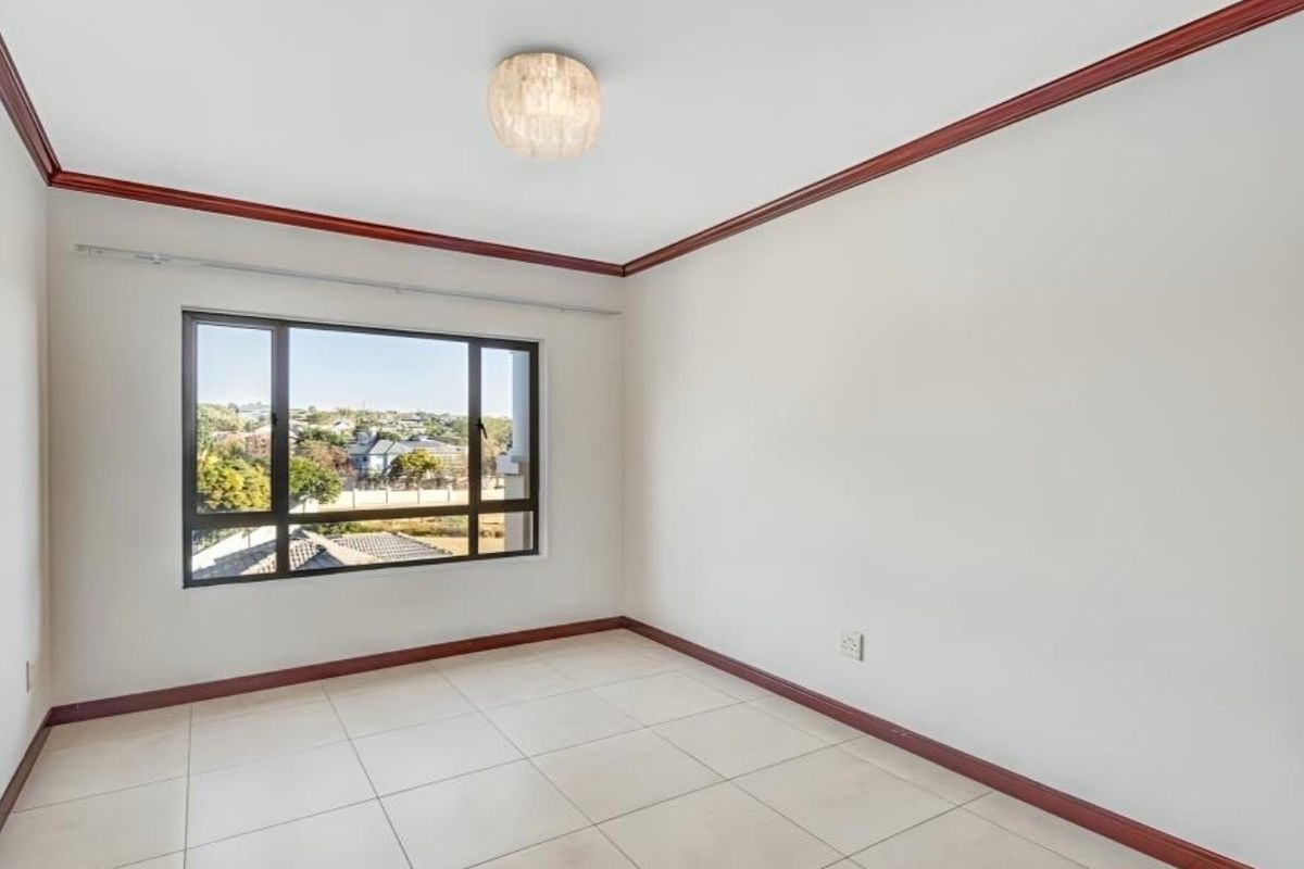 A bright, empty bedroom featuring neutral tiled flooring, crown molding, and a large window providing natural light and neighborhood views.