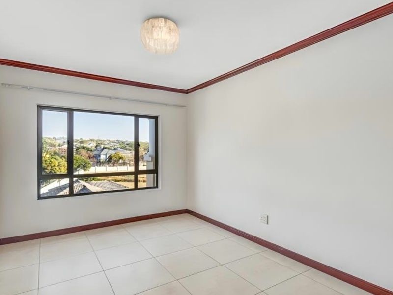 A bright, empty bedroom featuring neutral tiled flooring, crown molding, and a large window providing natural light and neighborhood views.