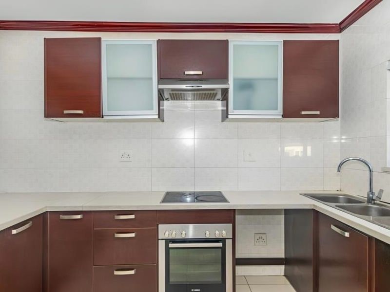 A clean, functional kitchen featuring dark wood cabinetry, stone countertops, built-in oven, electric cooktop, and a stainless steel sink with tiled backsplash.