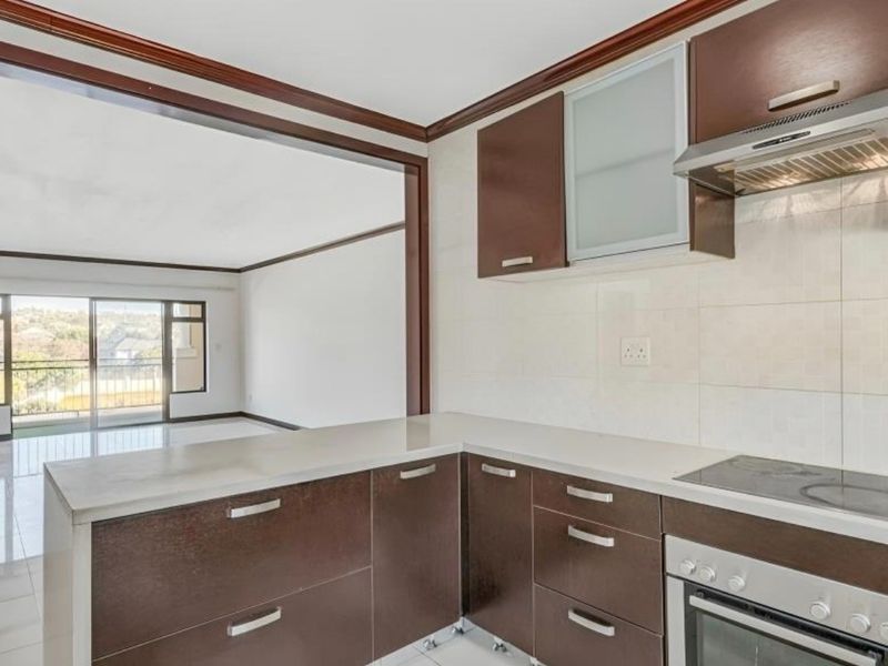 Open-plan kitchen featuring dark wood cabinetry, stone countertops, and integrated appliances, flowing into a bright living area with access to a balcony.