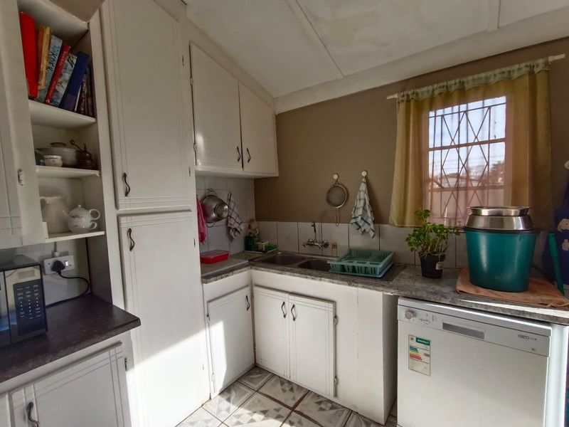 A compact kitchen featuring white built-in cabinetry, a double basin sink, tiled splashbacks, and tiled flooring with security bars on the window.