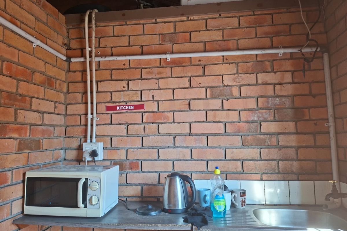 A basic kitchenette area featuring exposed brick walls, a stainless steel sink, and visible utility piping, suitable for a functional commercial workspace.