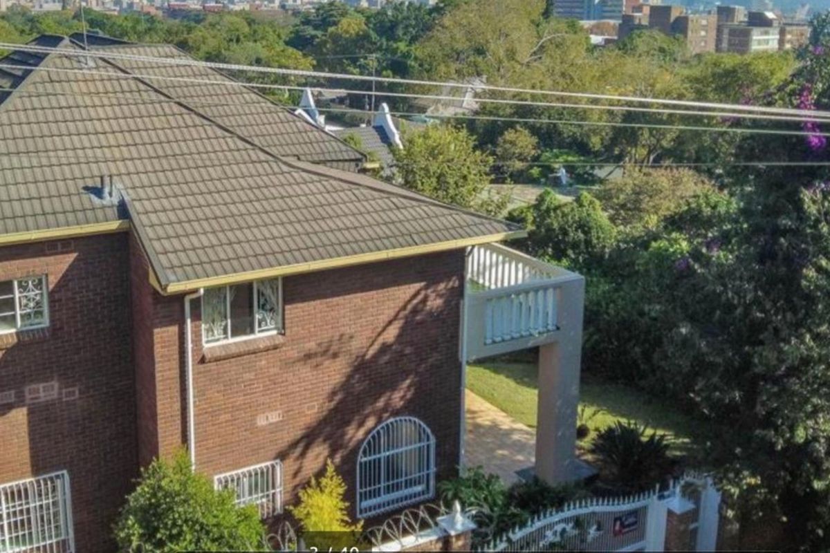 Elevated view of the rear exterior featuring a brick facade, tiled roof, and a balcony overlooking the surrounding garden and neighborhood.