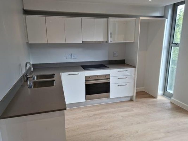 Modern kitchen featuring white cabinetry, stone countertops, integrated oven and hob, and light wood-look laminate flooring with natural light from a tall window.