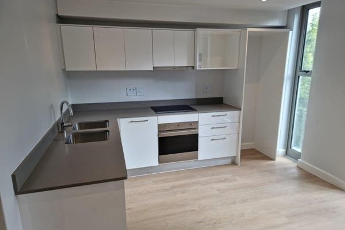Modern kitchen featuring white cabinetry, stone countertops, integrated oven and hob, and light wood-look laminate flooring with natural light from a tall window.