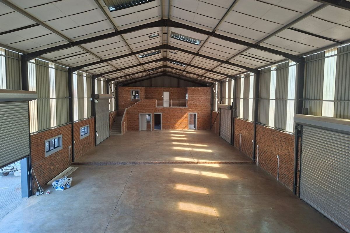 Spacious industrial warehouse featuring high ceilings with skylights, exposed brick walls, concrete flooring, multiple roller shutter doors, and an integrated mezzanine level.