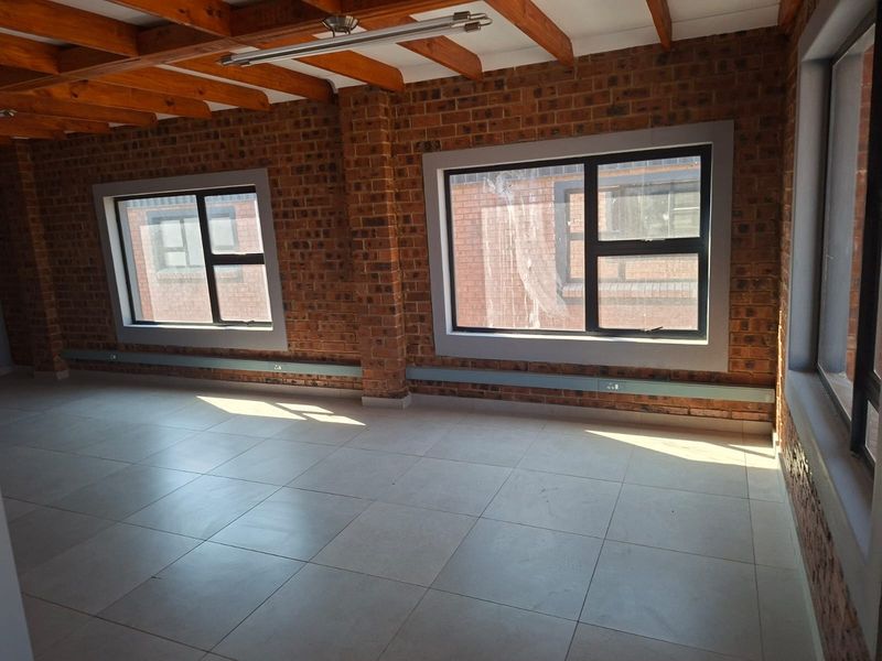 An open-plan office space featuring exposed brick walls, timber ceiling beams, tiled flooring, and large aluminium-framed windows providing natural light.