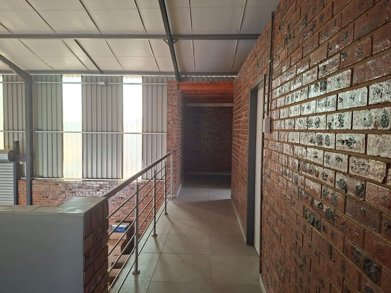 An industrial-style mezzanine hallway featuring exposed brick walls, tiled flooring, and a metal safety railing overlooking the lower level.