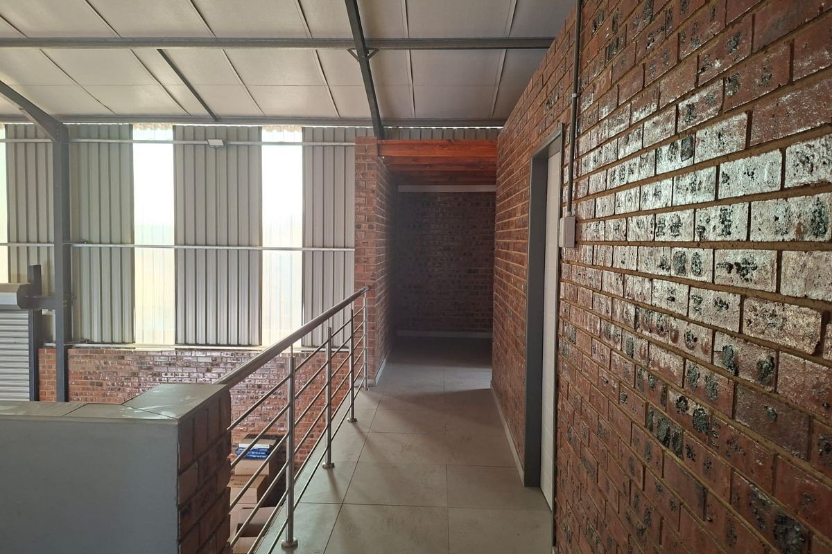 An industrial-style mezzanine hallway featuring exposed brick walls, tiled flooring, and a metal safety railing overlooking the lower level.