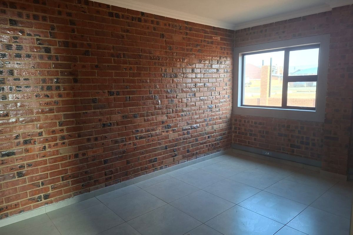An empty bedroom featuring exposed brick walls, neutral tiled flooring, and a large aluminium-framed window providing natural light.