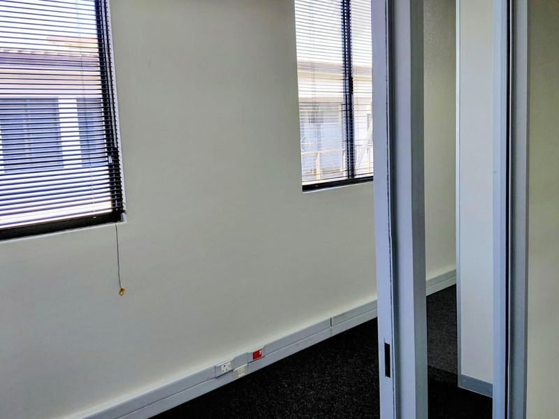 A clean, empty office space featuring dark carpeting, white walls, and venetian blinds on the windows, providing a functional professional environment.