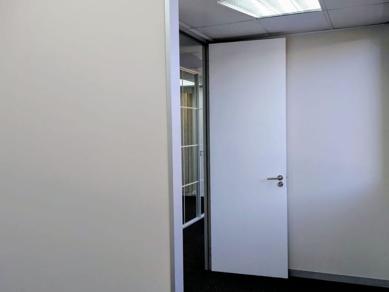 A clean, empty office space featuring neutral walls, carpet flooring, recessed ceiling lighting, and a glass partition door.