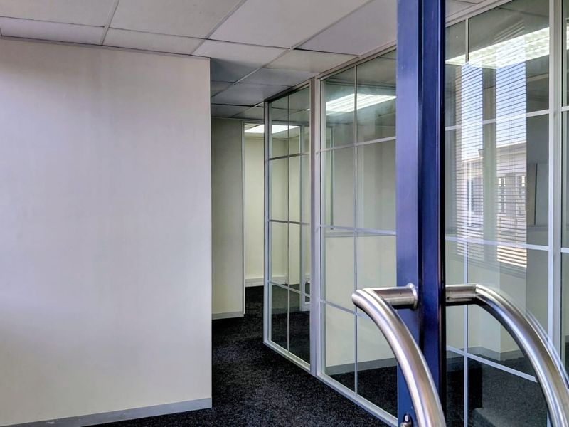 A vacant office space featuring glass partition walls, suspended ceiling tiles, and dark carpet flooring, offering a clean and professional layout.