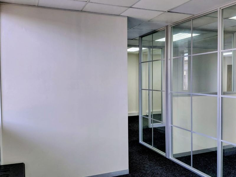 A vacant office space featuring glass partition walls, suspended ceilings with recessed lighting, and dark carpeting, offering a clean and professional layout.