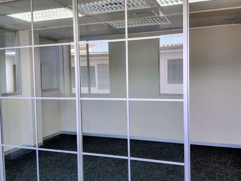 A bright, empty office space featuring a modern glass partition wall, suspended ceiling with recessed lighting, and dark carpet flooring.