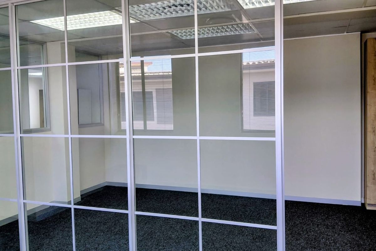 A bright, empty office space featuring a modern glass partition wall, suspended ceiling with recessed lighting, and dark carpet flooring.