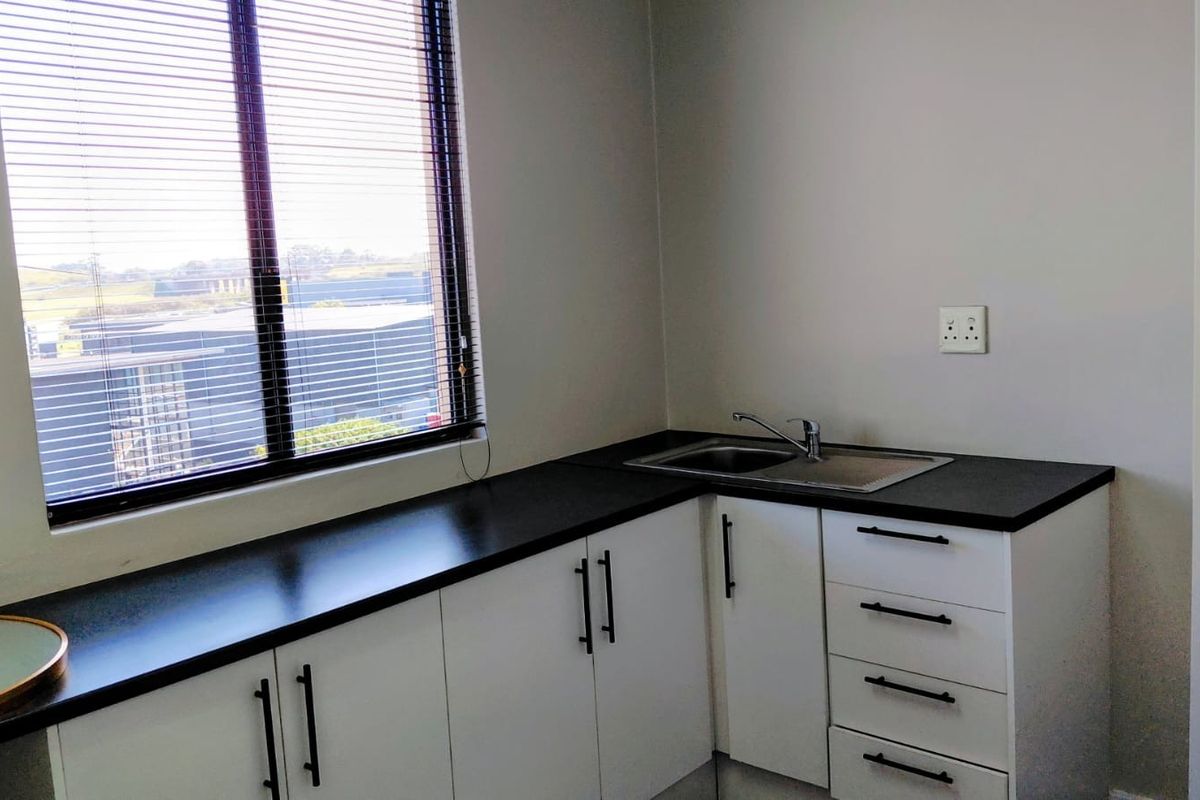 A clean, modern kitchenette featuring white cabinetry, dark laminate countertops, a stainless steel sink, and large windows with venetian blinds.
