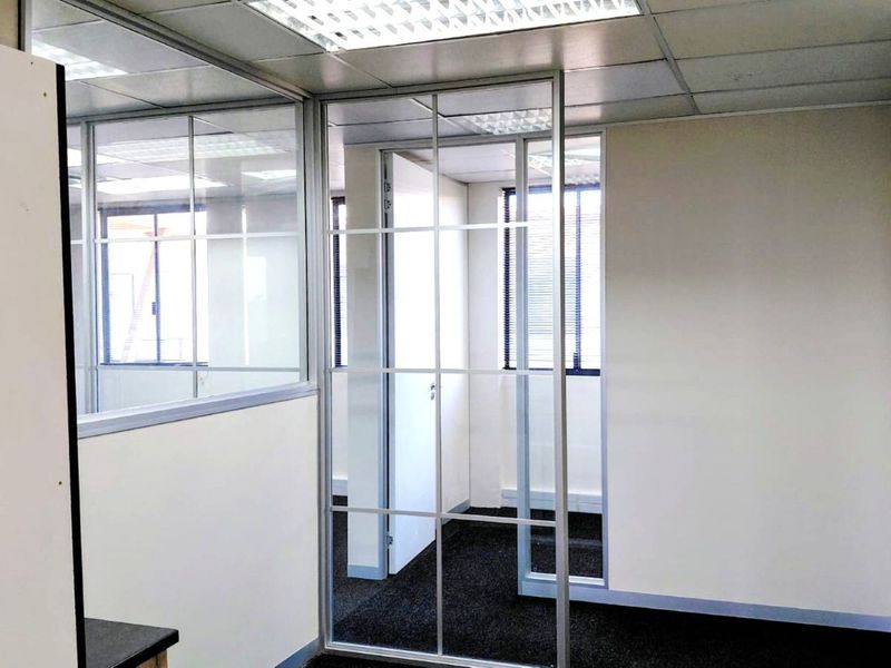 A vacant office space featuring glass partition walls, suspended ceilings with recessed lighting, and dark carpet flooring, offering a clean professional environment.