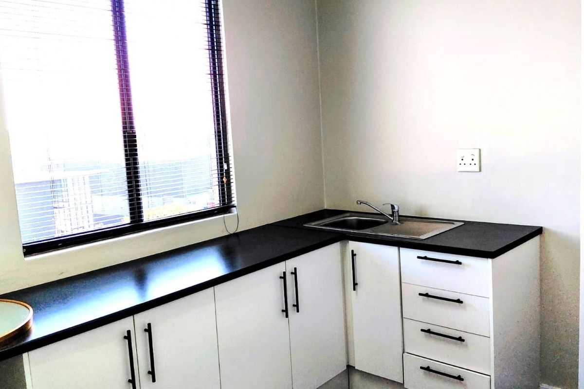 A compact, modern kitchenette featuring white cabinetry, dark countertops, a stainless steel sink, and a large window with venetian blinds.