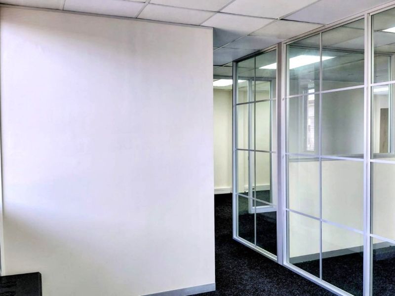 A bright, empty office space featuring modern glass partition walls, suspended ceilings, and dark carpet flooring, offering a clean and professional work environment.