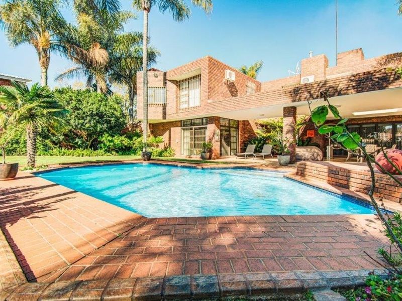 A spacious backyard featuring a swimming pool, brick-paved surround, and a covered patio area attached to the main brick residence.
