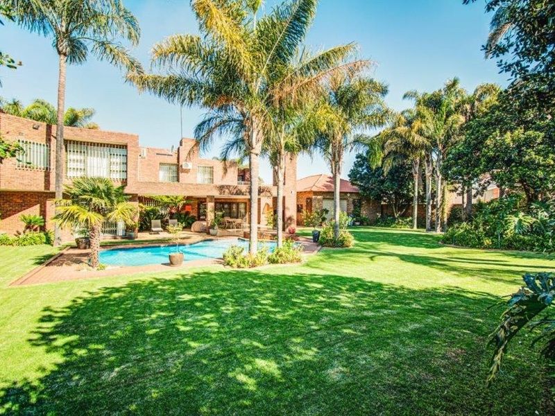 Spacious rear garden featuring a well-maintained swimming pool, lush lawn, mature palm trees, and a brick-faced residence with patio and balcony access.