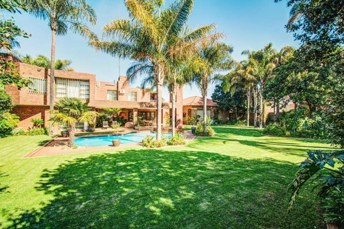 Spacious rear garden featuring a well-maintained swimming pool, lush lawn, mature palm trees, and a brick-faced residence with patio and balcony access.