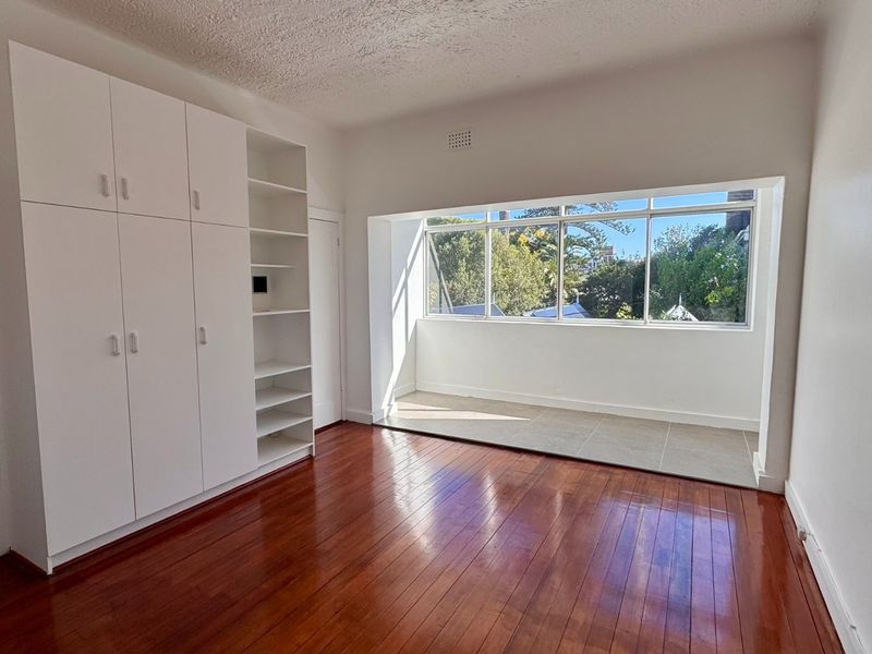 Bright bedroom featuring polished timber flooring, extensive built-in storage cabinetry, and a sun-drenched tiled alcove with large windows.