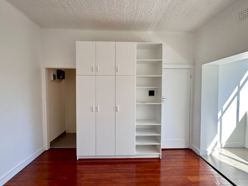 A bright, empty bedroom featuring polished hardwood flooring, ample built-in cupboard storage, and an integrated open shelving unit.