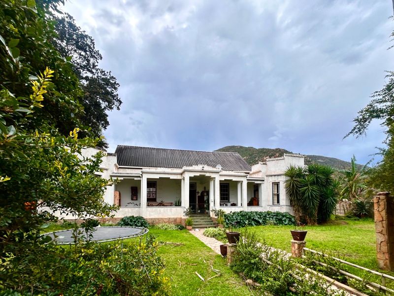 A charming farmhouse featuring a covered veranda, stone foundation, and a classic corrugated metal roof, set within a lush garden landscape.
