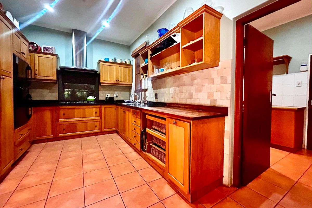 A traditional kitchen featuring warm wooden cabinetry, stone countertops, tiled flooring, and a built-in extractor fan, with an adjacent scullery area visible.