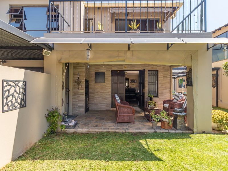 A covered patio area featuring tiled flooring, a built-in braai, and direct access to the garden, with an overhead balcony and railing.