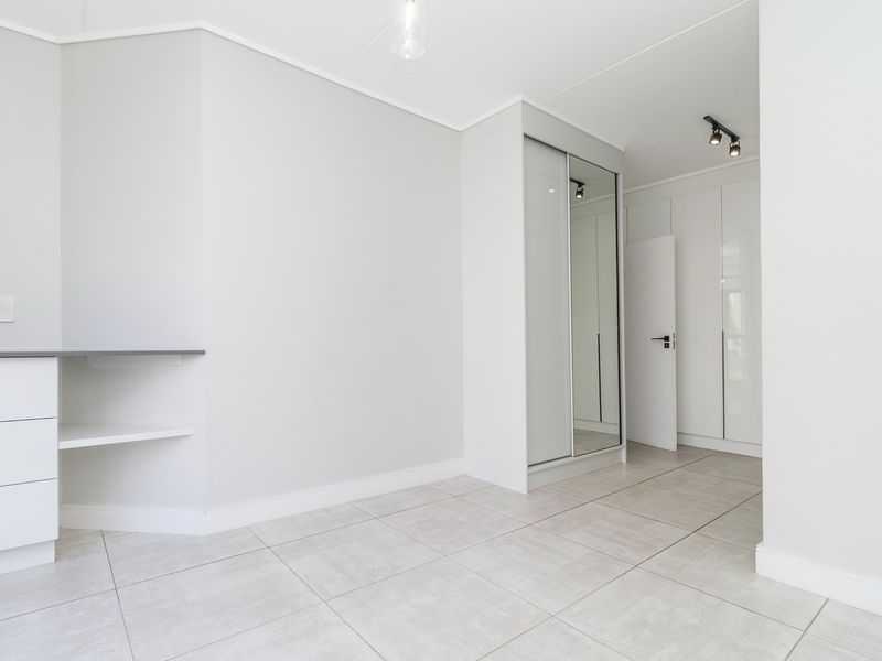 A bright, modern bedroom featuring neutral tiled flooring, ample built-in cupboard space with mirrored doors, and a convenient built-in desk area.