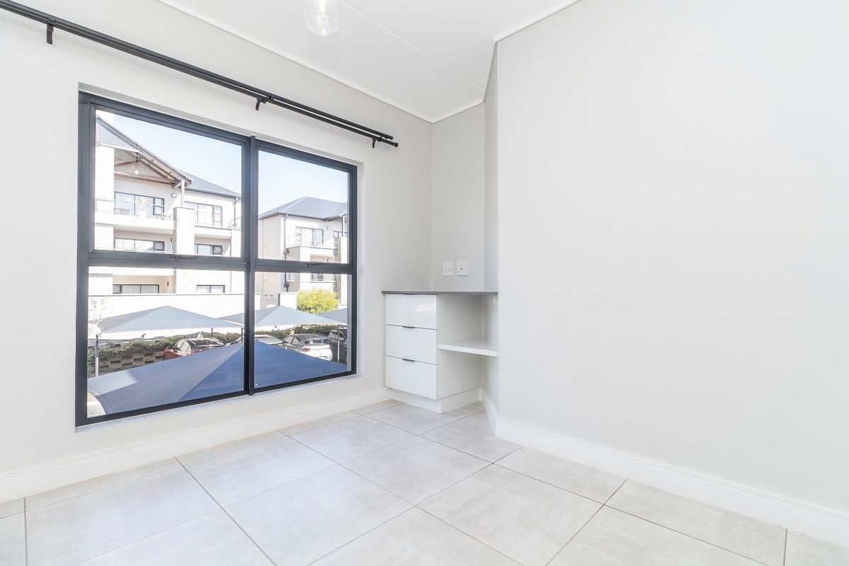 A bright, empty bedroom featuring neutral tiled flooring, a built-in desk unit, and large windows with views of the complex.