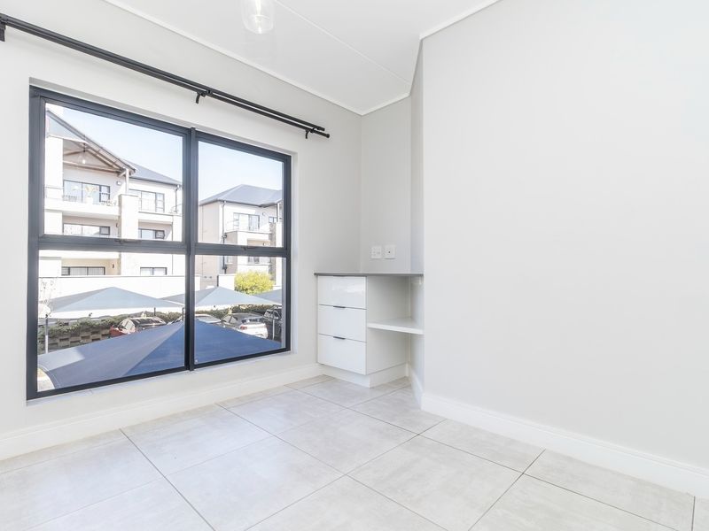 A bright, empty bedroom featuring neutral tiled flooring, a built-in desk unit, and large windows with views of the complex.