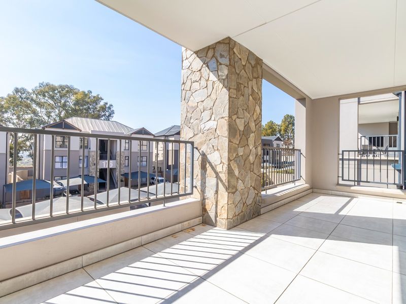 A spacious covered balcony featuring tiled flooring, a decorative stone-clad pillar, and metal balustrades, offering an open view of the apartment complex.