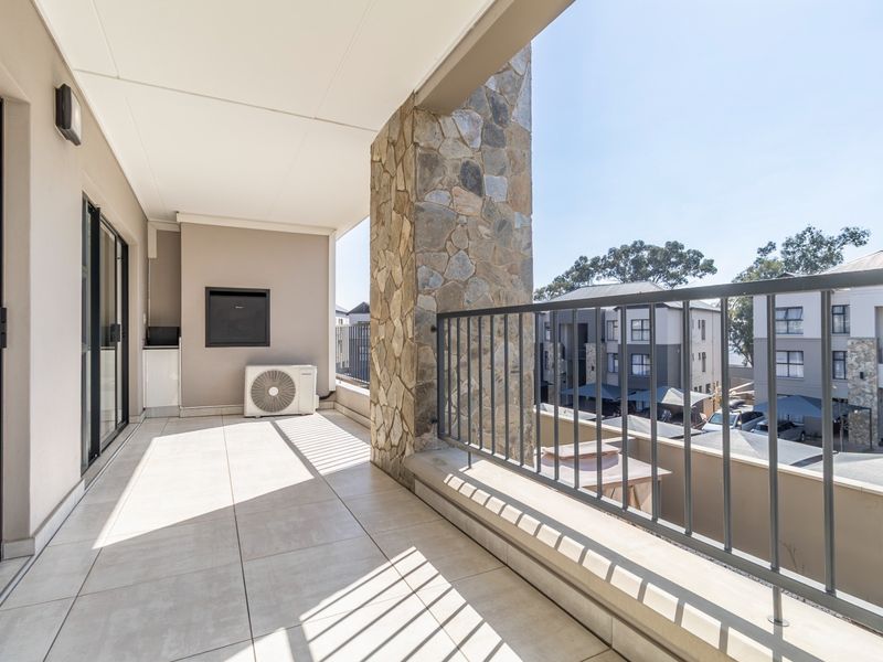 Spacious covered balcony featuring tiled flooring, a stone-clad pillar, built-in braai, and metal balustrade, offering a clean and well-maintained outdoor living space.