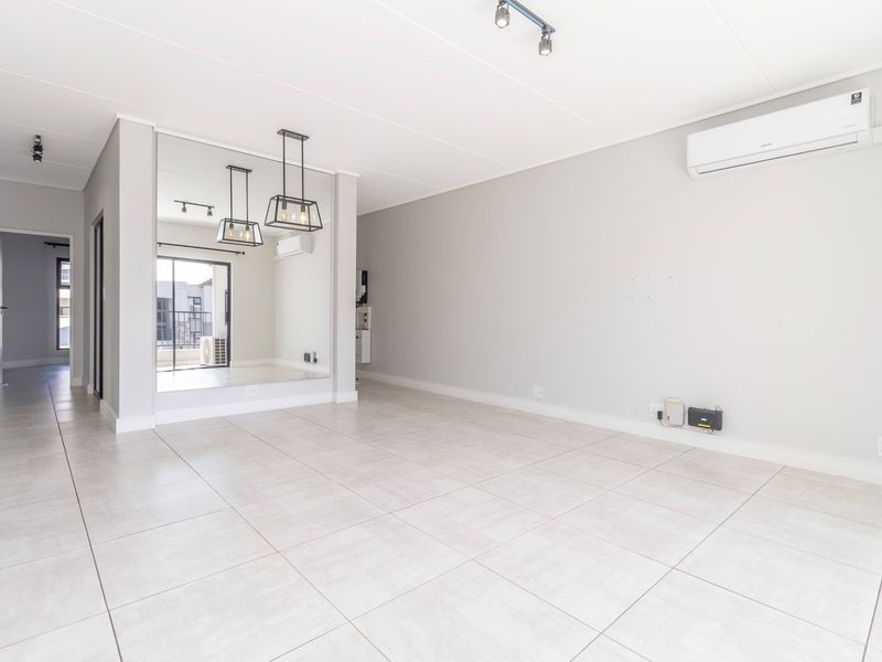 A bright, open-plan living area featuring neutral tiled flooring, a wall-mounted air conditioning unit, modern track lighting, and a large decorative mirrored wall.