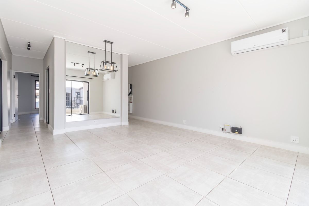 A bright, open-plan living area featuring neutral tiled flooring, a wall-mounted air conditioning unit, modern track lighting, and a large decorative mirrored wall.