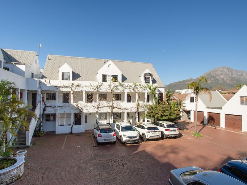Exterior view of an apartment complex featuring a paved courtyard, covered parking, and a multi-story building with a pitched roof.